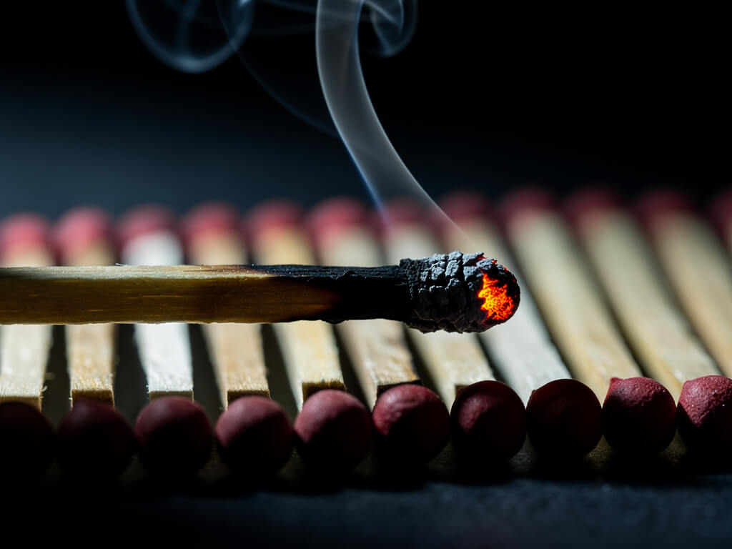 Single burnt matchstick still faintly smoldering next to a row of fresh unlit matches, dark background, dramatic macro photography, warm ember glow against cold tones