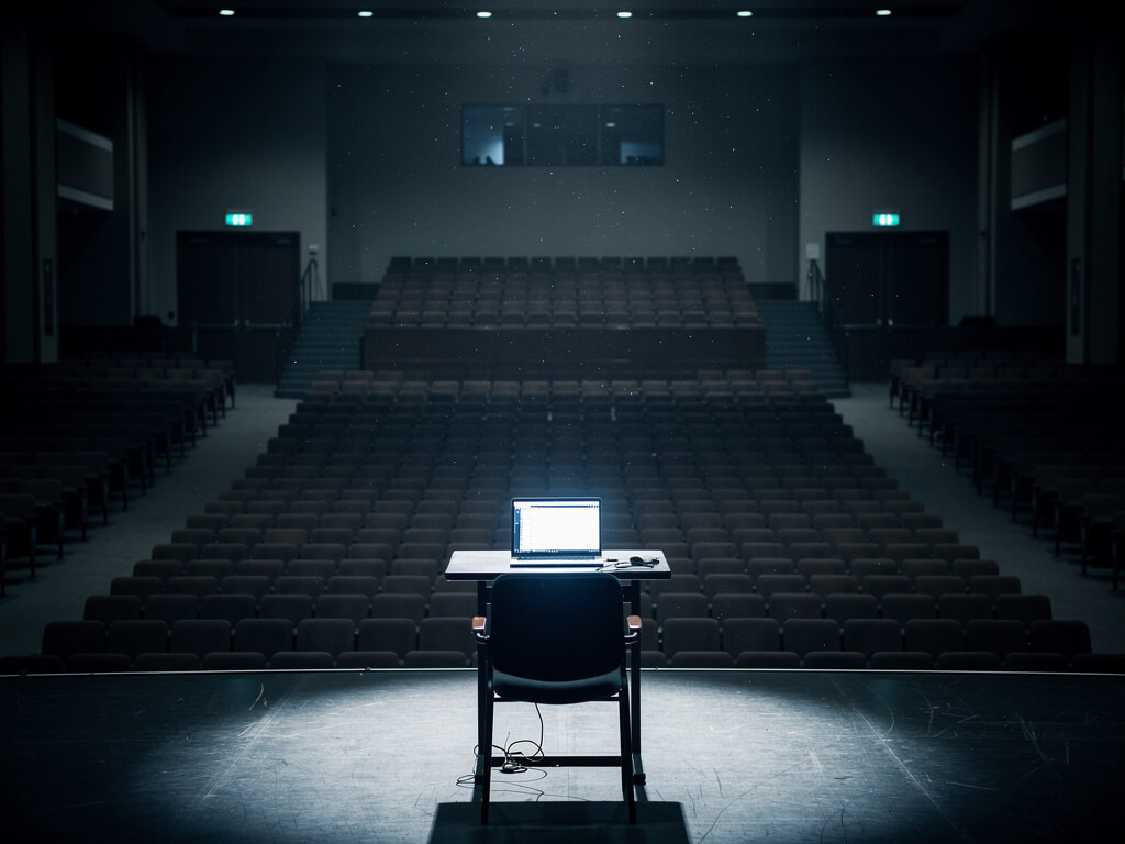A vast empty auditorium with hundreds of vacant seats, a single laptop glowing on stage presenting to nobody, hollow silence and wasted effort